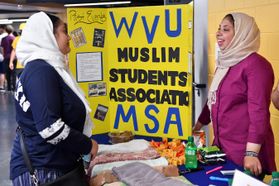A sign reads "WVU Muslim Students Association MSA" while two students in hijabs interact. 