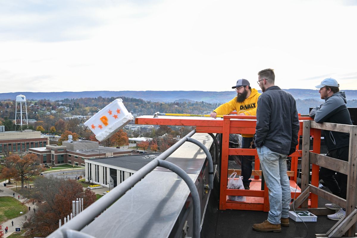 This photo is from the top of a building. It shows three people pushing a white package with a pumpkin on it off a bridge extending out over an 11-story drop.