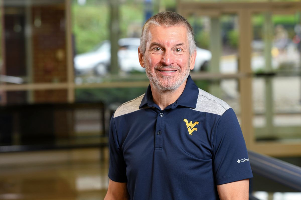 Photograph of Brad Humphreys, WVU professor. He is wearing a WVU branded, navy blue golf shirt and standing inside of a room full of widows in the background. He has short hair and a gray beard. 
