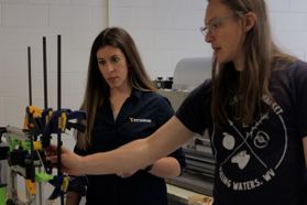 WVU researchers work together in a lab using specialized equipment. One woman is standing with her hand on a piece of equipment and speaking to a second woman standing next to her. 