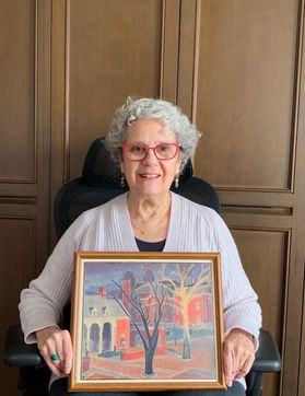 Photo of Suzanne Temple, former WVU student. She is pictured against a wall of wood paneling seated in a black chair. She is holding a colorful painting of WVU. She is wearing a light colored cardigan with a black shirt underneath. She has curly gray hair