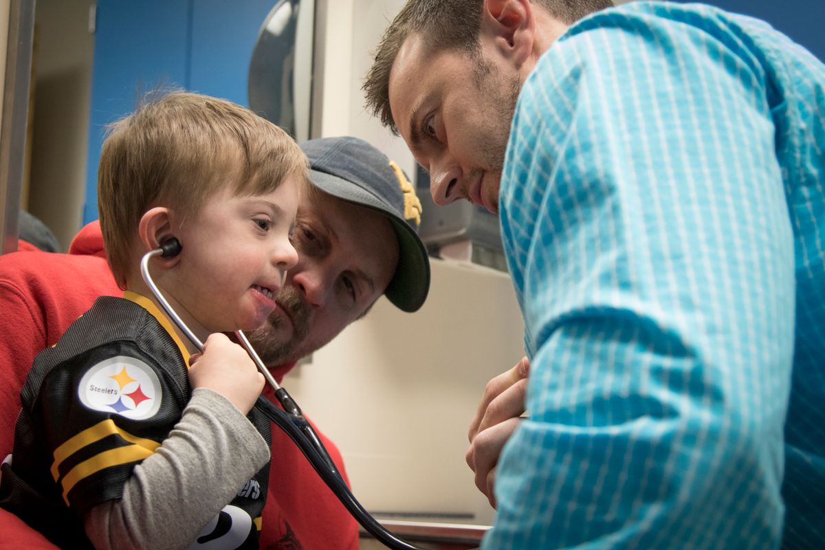 A child tries out a stethoscope while sitting on an adult's lap. Another man is also in the photo.