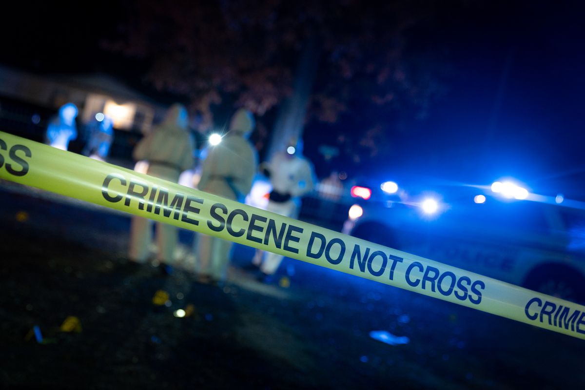 An image depicting a crime scene with crime scene tape in the foreground, police lights and cars in the background, and workers wearing yellow suits and black gloves standing near the cars.