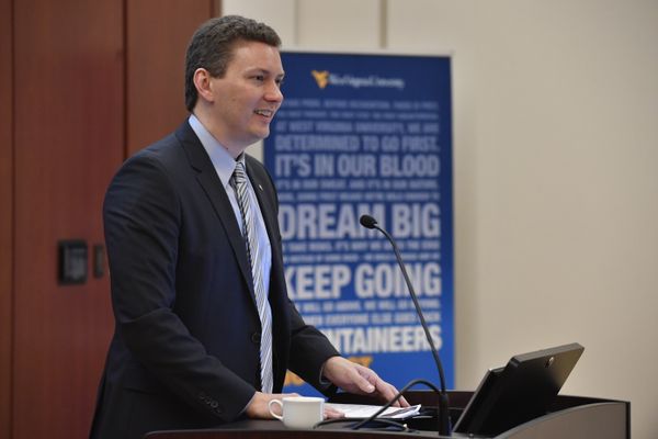 John Deskins, director at the WVU Bureau of Business and Economic Research, stands and smiles at a podium while wearing a blue shirt and tie with a black jacket.