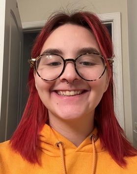 Headshot of WVU student Red Klug. They are pictured inside with a door frame in the background. They are wearing an orange hoodie and have bright pink hair with round framed glasses. 
