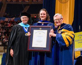 There people wear Commencement regalia. In the center, the 2023 Presidential Honorary Degree recipient holds a larger honorary degree.