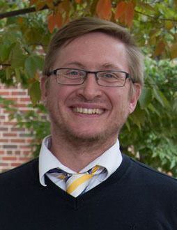 Headshot of Sam Taylor. He is pictured outside with a brick wall and green tree just behind him. He is wearing a navy blue v-neck sweater with a gold and blue striped tie. He has short blond hair, a short beard and wears square glasses. 