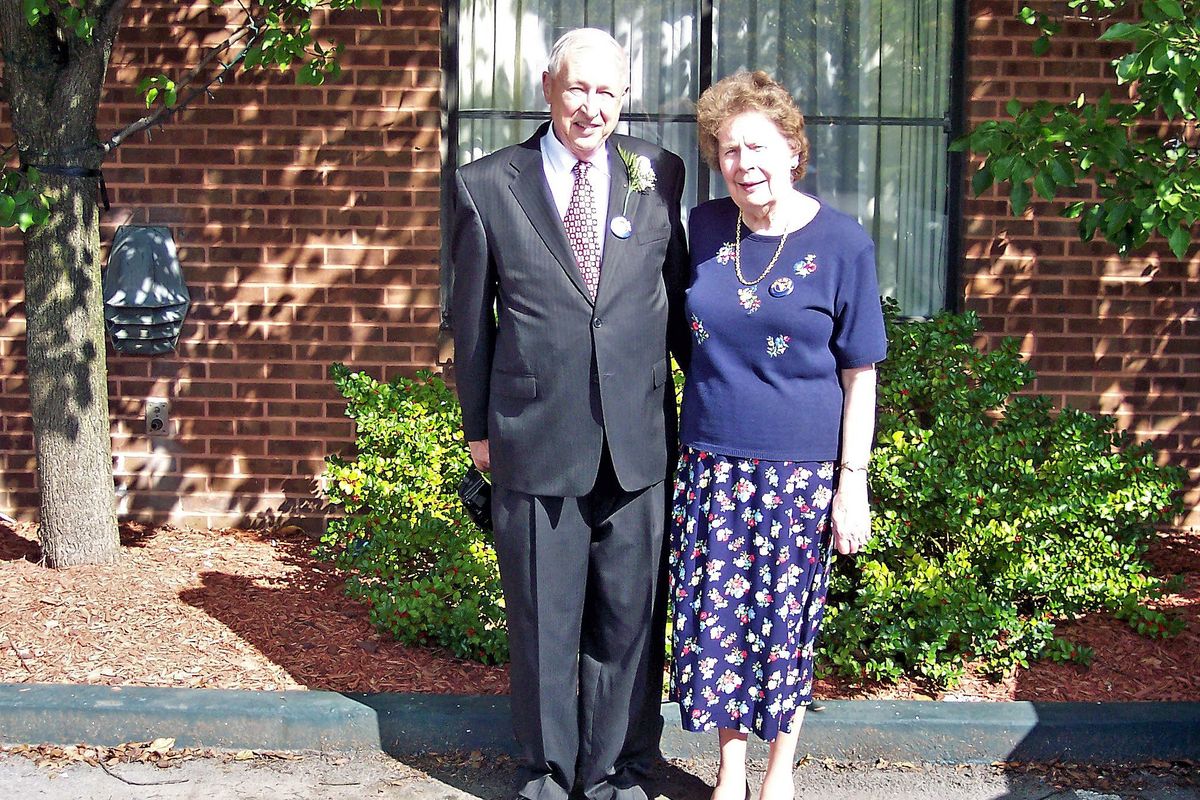 man in suit and tie, woman in sweater/skirt combo in front of brick wall, window