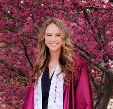 Scholarship recipient Taylor Kennedy is pictured standing in front of a tree full of bright pink flowers wearing a maroon graduation gown and a white "Student Athlete" sash.
