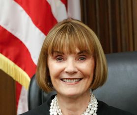 photo of smiling woman, chin-length hair, bangs, big white necklace, wearing black robe, sitting in black chair in front of a flag