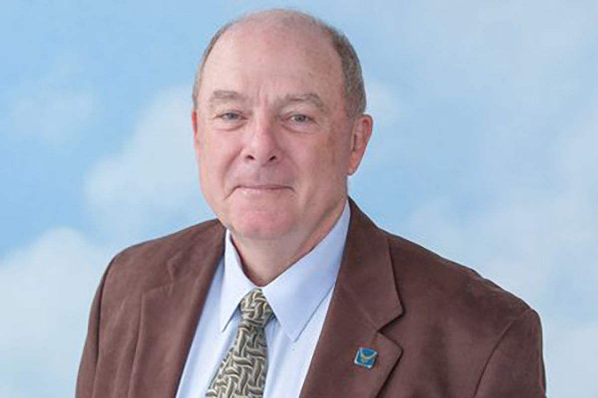 Photo of WVU alum. He is an older man pictured in front a light blue background with white puffy clouds. He is wearing a brown jacket over a light blue dress shirt and patterned tie. 
