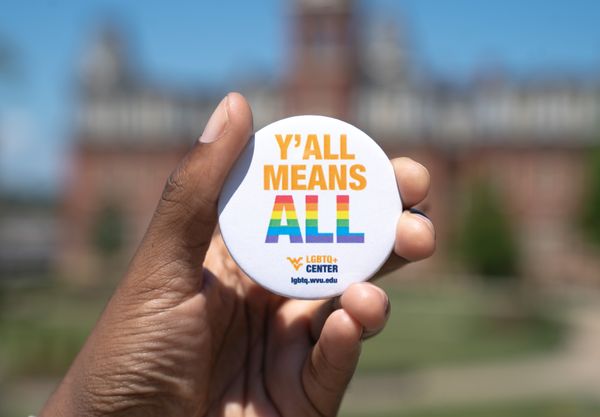 A hand is visible holding a white button that says 'Y'all means ALL.' On the button ALL is in rainbow colors. Woodburn Hall is blurry in the background.