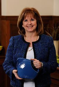 Elizabeth McIlvain is pictured here wearing a royal blue suite that perfectly matches the royal blue Fiestaware pitcher she is holding. Her strawberry blond hair is shoulder length.