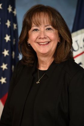 Headshot of Karen Evans. She is pictured against a patriotic background with an American flag on her left. She is wearing a black suit and has shoulder-length auburn hair with bangs. 