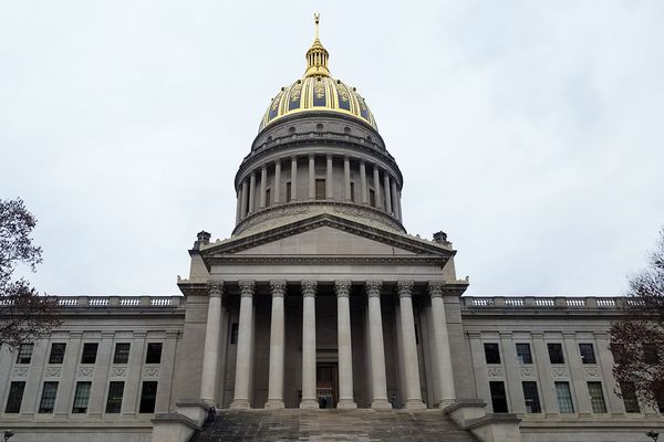 This is a photo looking up at the West Virginia State Capitol from the side that faces the Kanawha River. In the middle of the frame are columns. The gold and blue dome is against a gray sky.