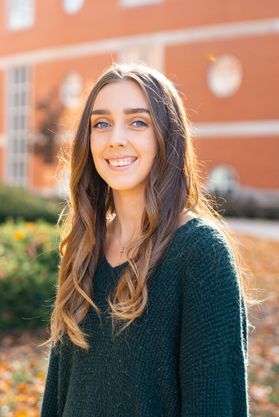 Photograph of scholarship recipient Madison Branham. She is pictured outside with a red brick building behind her. She is wearing a deep green sweater and has long, light brown hair. 