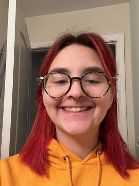 Headshot of WVU student Riley Klug. She is pictured inside and is wearing an orange sweatshirt. She has dyed red hair and wears tortoise shell round glasses. 