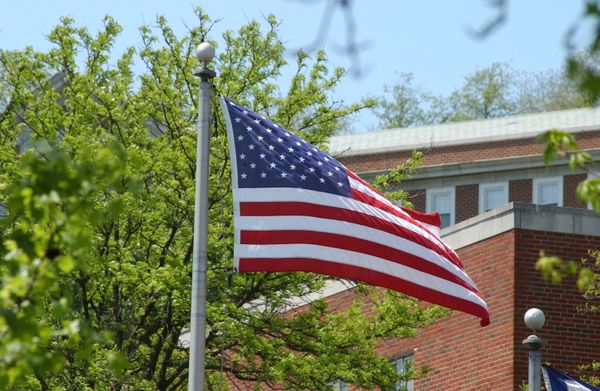An image of an American Flag on a flagpole in front of a red brick building. There are green trees behind the flag pole.
