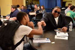 Man wearing black shirt and sunglasses fist bumps student wearing white shirt and black backpack while sitting at a desk
