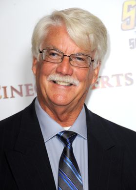 Photo of smiling man with white hair and mustache wearing dark suit jacket, blue shirt, blue and black striped tie, glasses