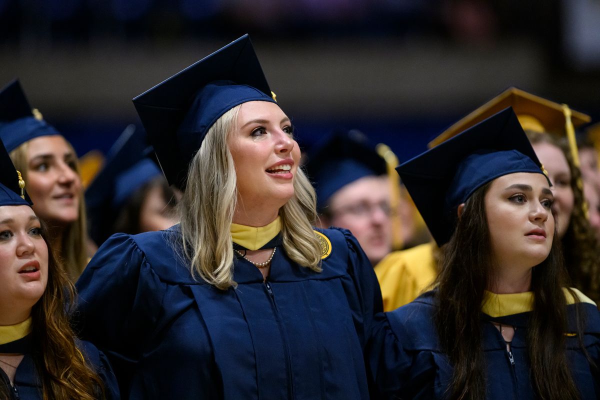 WVU graduates are shown  standing in rows in their navy blue and gold regalia during a Commencement ceremony. 