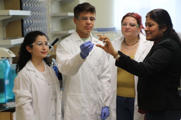 Professor Soumya Srivastava works with three students in her lab on a project to create tick-borne disease testing. Two student are female and one is male. All three are wearing lab coats. The professor is dressed in black. 