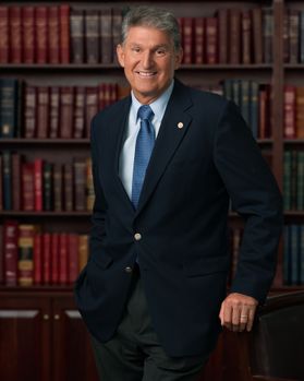 Headshot of U.S. Sen. Joe Manchin standing in front of stacks of legal books with one hand rested on the back of a chair. He is wearing a navy blue suit over a white dress shirt with a light blue colored tie. He has short, gray hair. 
