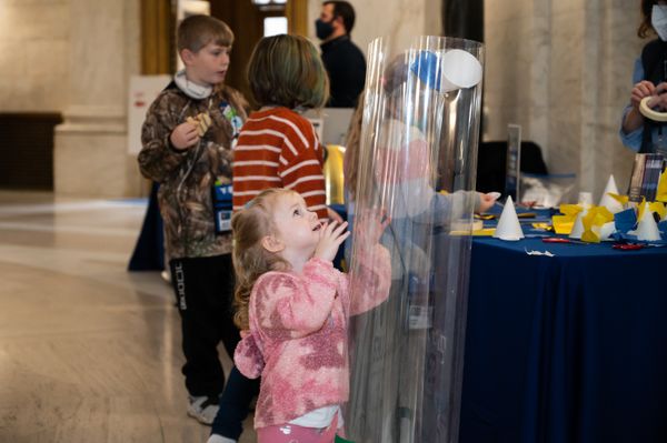 This photo features a young girl dressed in pink with blond hair examining a clear plastic tube that is taller than her at the WVU Day at the Legislature. Other kids are shown in the background visiting tables and display in the Capitol rotunda. 