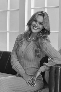 Headshot of WVU student Isabella Crouch. The photo is black and white and shows Crouch seated on a leather couch. She has long blond hair and is wearing a ribbed sweater dress. 