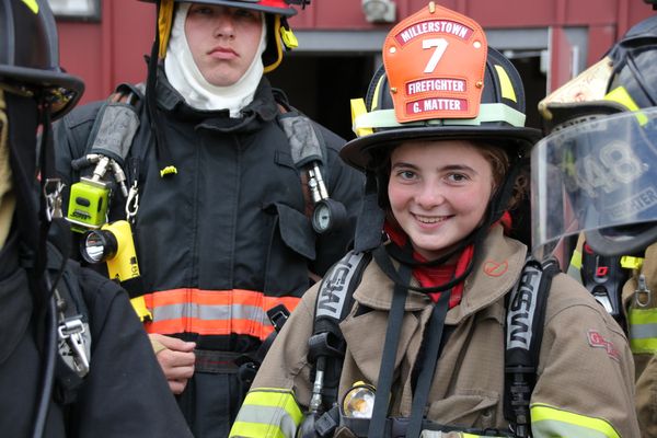 A young person smiles at the camera while wearing full firefighting gear, including a hat. Parts of three other people in firefighting gear are in the frame as well.