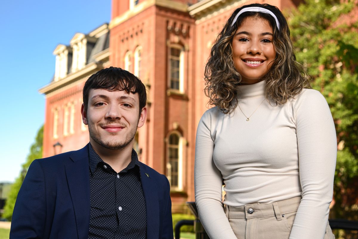 Ariana wears a headband and light color short with dress pants stands next to Matthew who wears a dress shirt with suit jacket in front of Woodburn Hall.