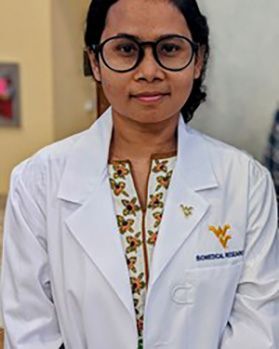 Headshot of WVU graduate student Yeasmin Akter. She is standing inside wearing a white, WVU lab coat over a yellow and off white floral top. She has dark hair pulled back and wears glasses. 