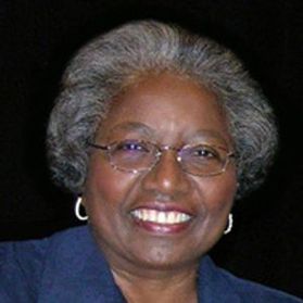 Headshot of Carolyn Bailey Lewis. She is pictured against a black background. She is wearing a blue top and has short black and gray hair. She is also wearing oval-shaped glasses. 