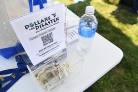 A clear box with money sits on a white table. Over the clear box is a paper that reads, 'Dollars for Disaster.' A water bottle is also on the table to the right of the box.
