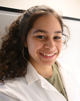 Headshot of WVU student Rachel Morris. She is pictured wearing a white lab coat over an olive colored shirt. She has long brown curly hair. 