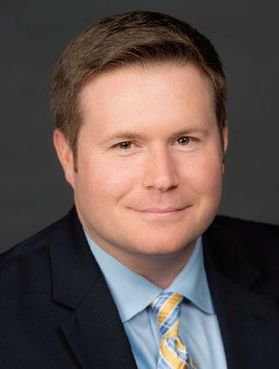 Headshot of WVU administrator Travis Mollohan. He is pictured against a dark gray background wearing a dark suit jacket over a light blue shirt and a blue and gold patterned tie. He has short light colored hair.  