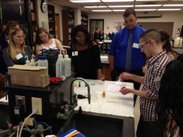 five students wearing lab goggles stand at white desk with adult in blue shirt and black tie 