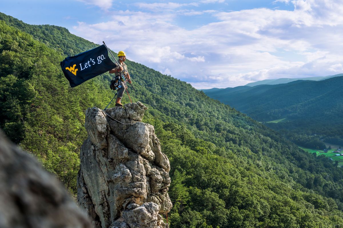 A mountaineer stands at point of a rocky cliff, rolling hills and a cloudy sky behind him. He holds a West Virginia University 