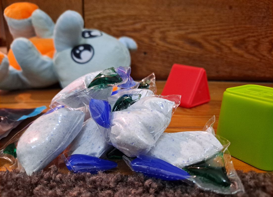 Image of common household detergent pods laying on the ground near children's toys including a green and a red building block, and a gray and orange stuffed elephant. 