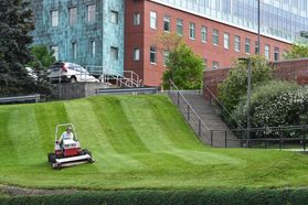 man on large mower cuts grass in front of large building in a hillside