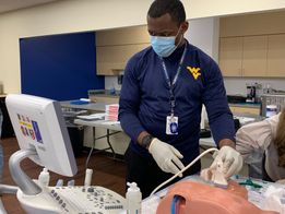 A Black WVU Health Sciences student wearing a navy blue shirt with a Flying WV on the left pocket, a light blue surgical mask and gloves, is shown here in a simulation lab working with ultrasound equipment and a dummy. 