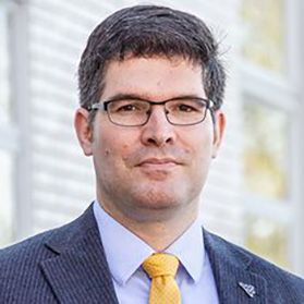 Headshot of WVU researcher Thorsten Wuest. He is pictured with a blurred building behind him. He is wearing a navy blue sports coat over a light blue dress shirt and a gold tie. He has short dark hair and wears glasses. 