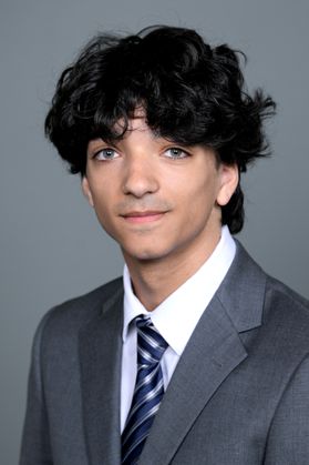 Headshot of WVU Bucklew Scholar Alexander Tadros. He is pictured against a gray background wearing a gray suit with a white dress shirt and blue and silver striped tie. He has medium length curly dark hair. 