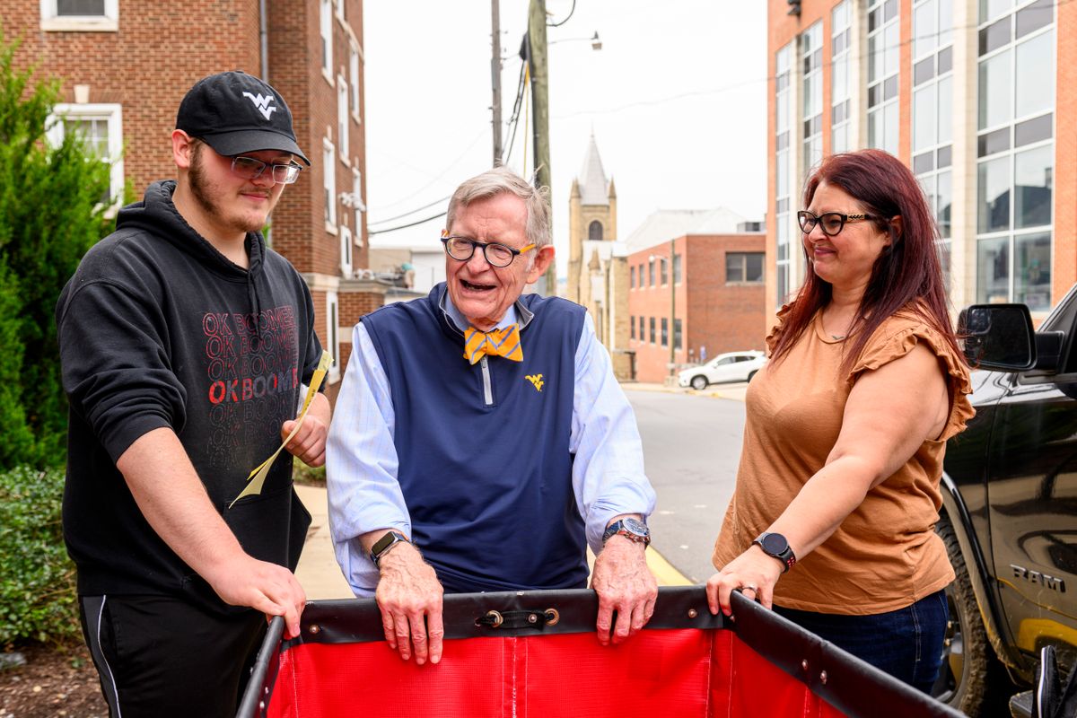 WVU President Gordon Gee talks to new students moving on to the WVU Campus. He is standing between the two of them with his hands resting on a red move-in bin. 