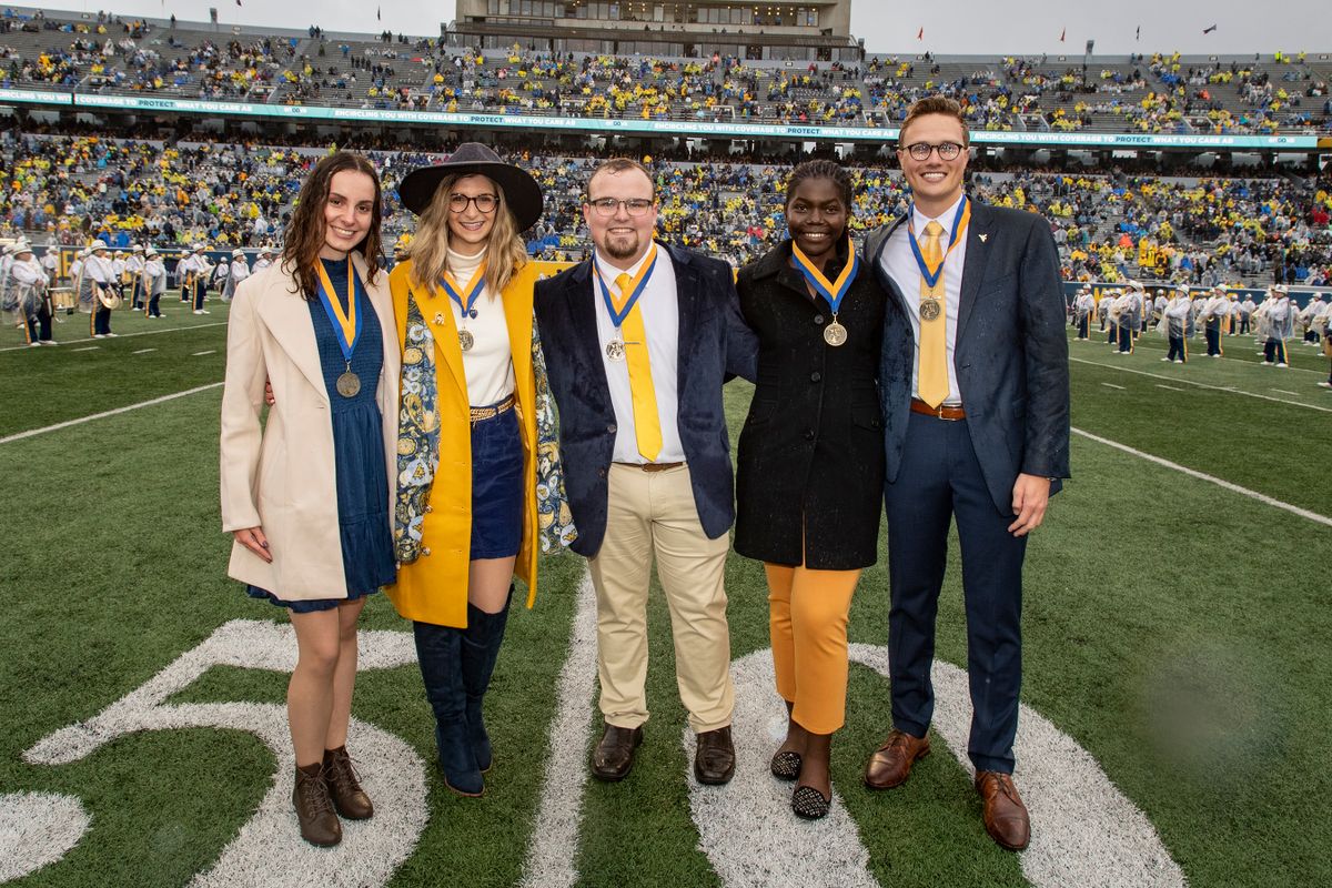 The five winners of the Mountaineers of Distinction awards stand on Mountaineer Field in the rain. They are wearing colors of gold and blue. Fans are visible in the stands.