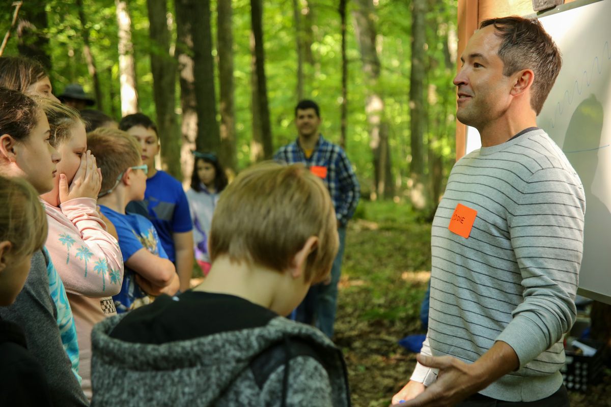 WVU biology professor Edward Brzostek teaches elementary students about forest ecosystems during a field trip into the woods. Brzostek is standing in front of the group wearing a gray striped seater talking to several grade schoolers. 