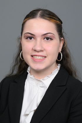 Headshot of WVU Foundation Scholar Mariana Alkhouri. She is pictured against a gray background and is wearing a black jacket and a white dress shirt with a ruffled neckline. She is wearing hoop earrings and has long, curly dark hair pulled up on the sides