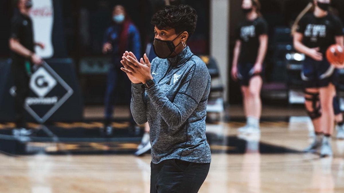 A woman with short dark hair in a gray sweatshirt and black mask claps while standing on a basketball court