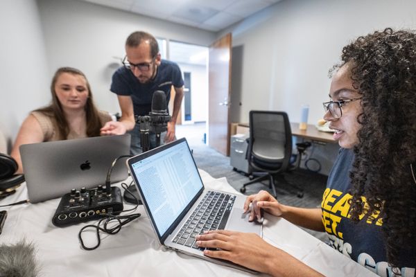 Three WVU researchers work together in a computer lab. One male researchers stands near a female researcher pointing at her computer screen. The third researcher sits near them working on her laptop. 