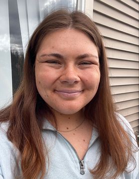 Headshot of WVU student Rachel Lasky standing in front of a building wearing a gray zip-up sweatshirt. She has long, light brown hair. 